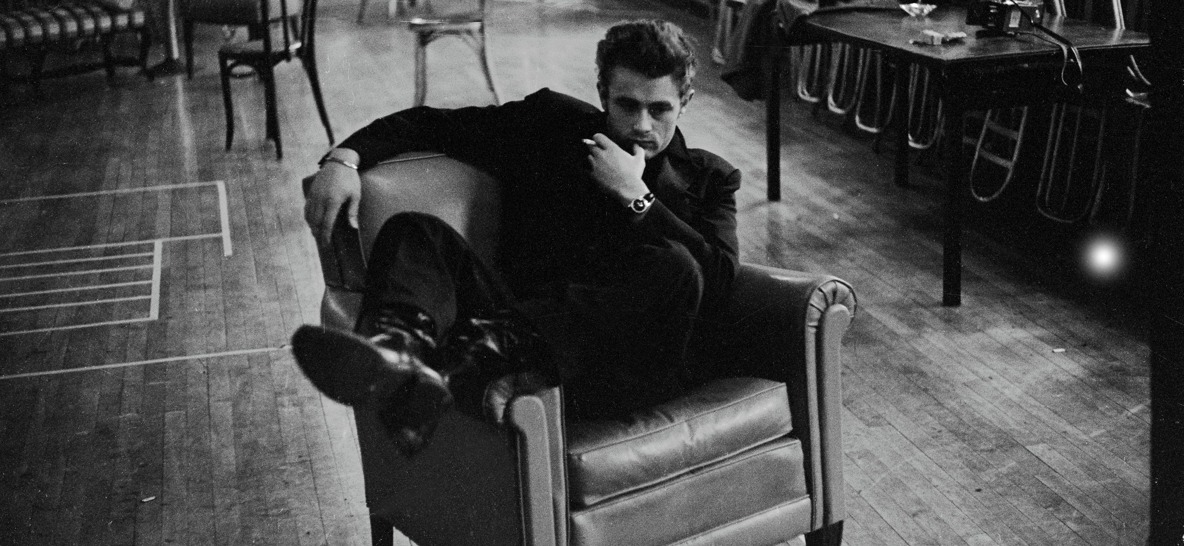 James Dean sits sideways in an arm chair, smoking a cigarette and thinking deeply.